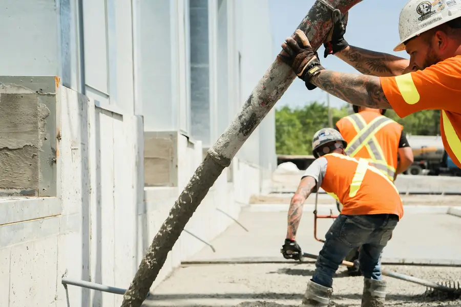 Two workers in high-visibility vests guiding a hose along a foundation wall